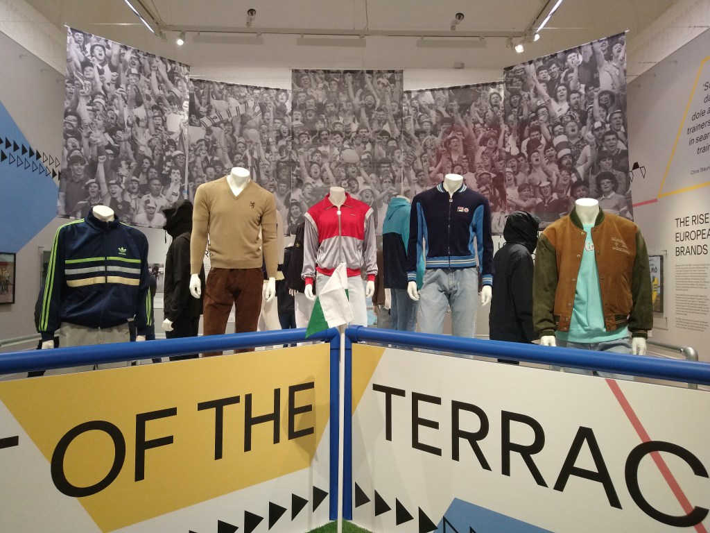 Image of various Mannequins on a terraced display. They are wearing various forms of Casual style fashions. Hung behind are images of football crowds. 